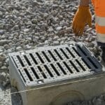 A worker wearing orange gloves stands near a newly installed concrete drainage grate on a bed of crushed stone.