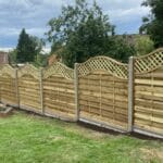 A newly installed wooden fence with lattice-top panels, neatly aligned in a green garden with trees and shrubs in the background.