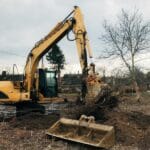 A large yellow excavator digging out tree roots on a construction site, preparing the land for development.
