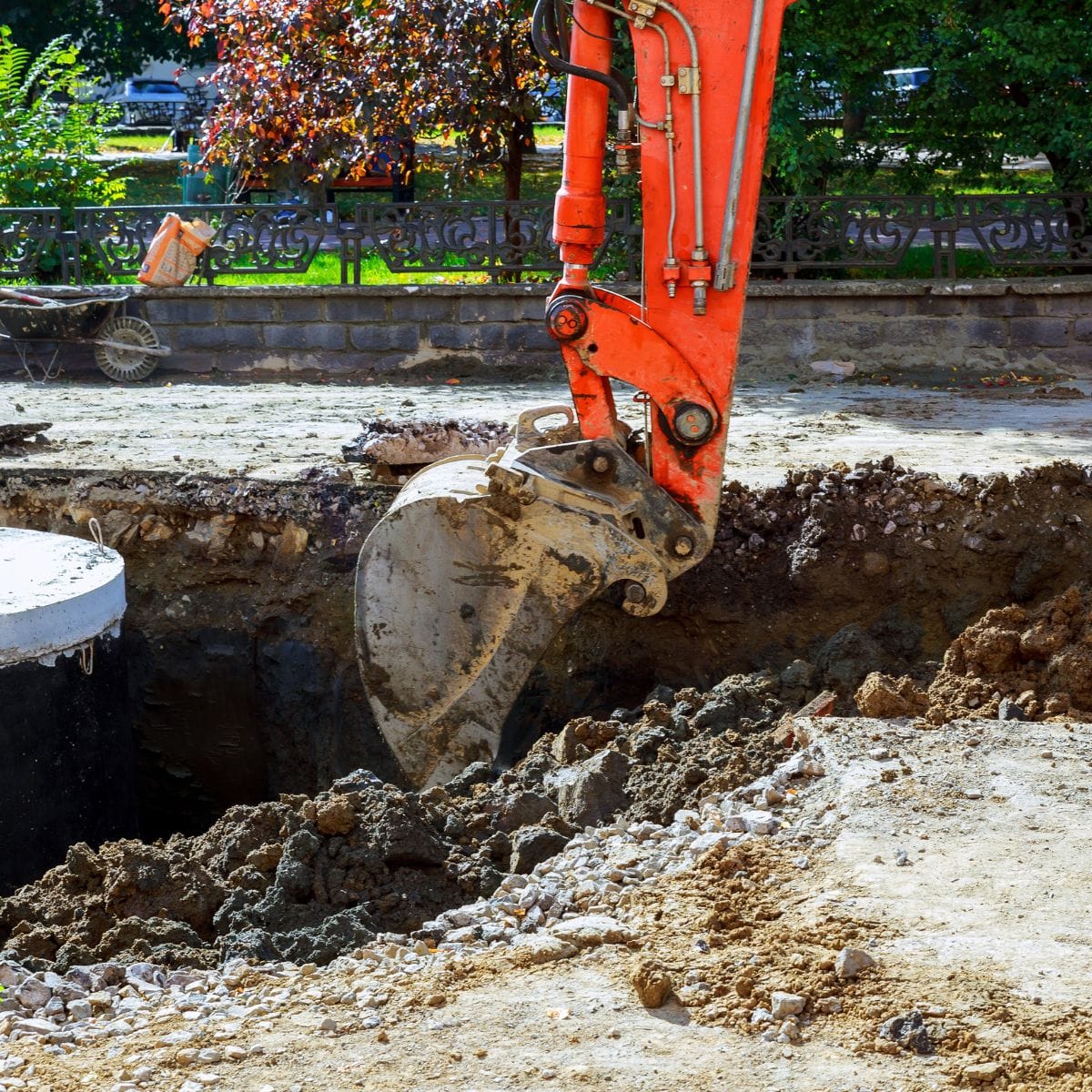 A close-up of an orange excavator digging a deep trench, with its bucket covered in soil and rubble.